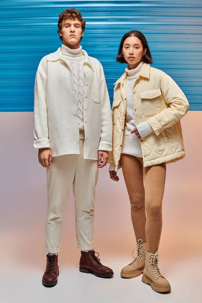Casal interracial em jaquetas quentes e botas de couro olhando para a câmera perto do painel de plástico azul — Fotografia de Stock