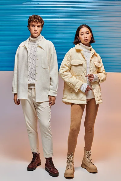 Comprimento total do casal interracial em jaquetas quentes e botas de couro posando perto de folha de plástico azul — Fotografia de Stock
