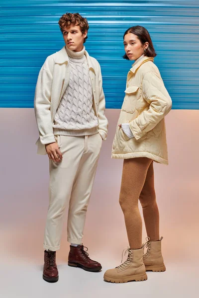Міжрасова пара в модному зимовому вбранні позує біля блакитного пластикового листа, сезонна мода — стокове фото