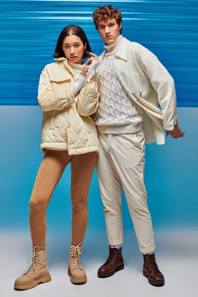 Lookbook de mode d'hiver, couple interracial à la mode regardant la caméra sur fond bleu avec du plastique — Photo de stock
