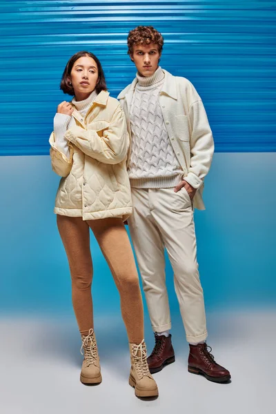 Trendiger Mann mit Hand in Tasche neben asiatischer Frau in warmer Kleidung auf blauem Hintergrund mit Kunststoffplatte — Stockfoto