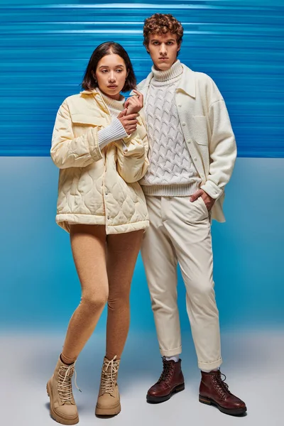 Стильный мужчина с рукой в кармане возле азиатской женщины в зимнюю одежду на синем фоне с пластиковым полотном — стоковое фото