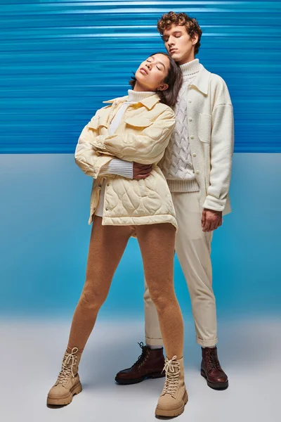 Asian woman in winter attire posing with closed eyes near stylish man on blue plastic background — Stock Photo