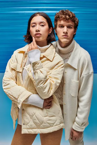 Campanha de moda de inverno, casacos de casal interracial olhando para a câmera no fundo de plástico azul — Fotografia de Stock