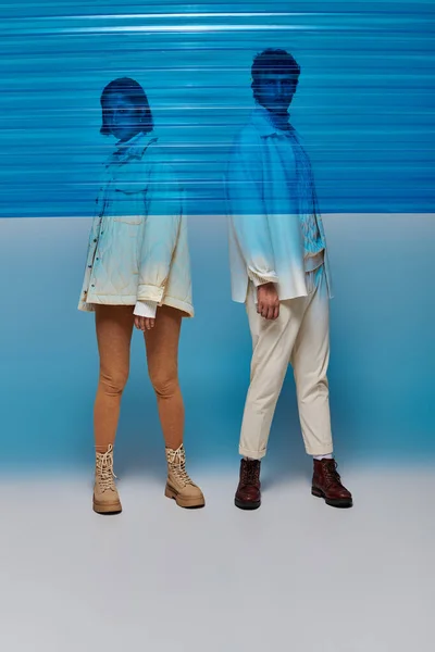 Multiethnisches Paar in Jacken und Lederstiefeln posiert hinter blauem Plastiktuch, Wintermode — Stockfoto