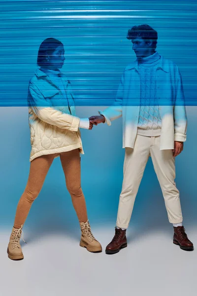 Volle Länge des stilvollen gemischtrassigen Paares, das die Hände hinter blauem Plastik im Studio hält, im Winteroutfit — Stockfoto