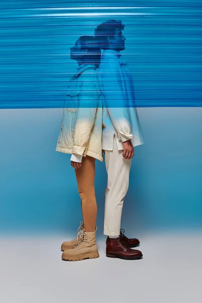 Vista lateral do casal interracial em roupas quentes de pé de volta para trás atrás de plástico azul em estúdio — Fotografia de Stock