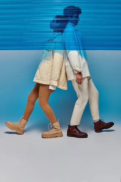 Vista lateral de modelos interracial en ropa de invierno posando espalda con espalda detrás de una lámina de plástico azul - foto de stock