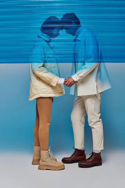 Casal interracial em roupas de inverno de pé cabeça a cabeça e de mãos dadas atrás de plástico azul — Fotografia de Stock