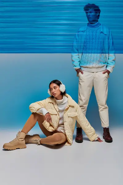 Модная азиатская модель в наушниках, сидящая рядом с мужчиной с руками в карманах и голубым пластиковым листом — стоковое фото