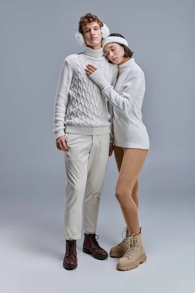 Азиатка в белом свитере и повязке, опирающаяся на грудь модного мужчины на серой, зимней моде — стоковое фото
