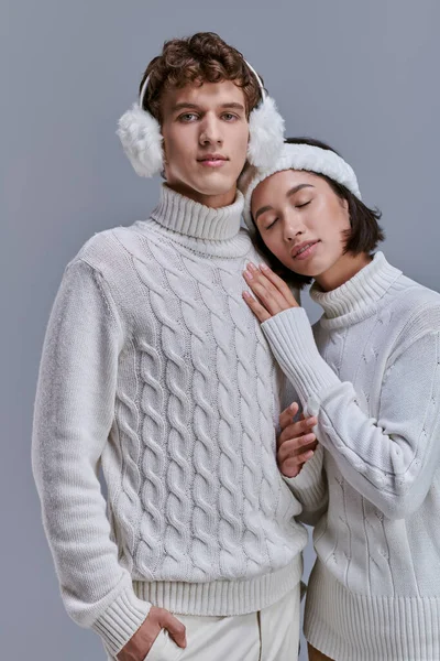 Felice donna asiatica in maglione bianco e fascia appoggiata sul petto dell'uomo in caldo paraorecchie su grigio — Foto stock