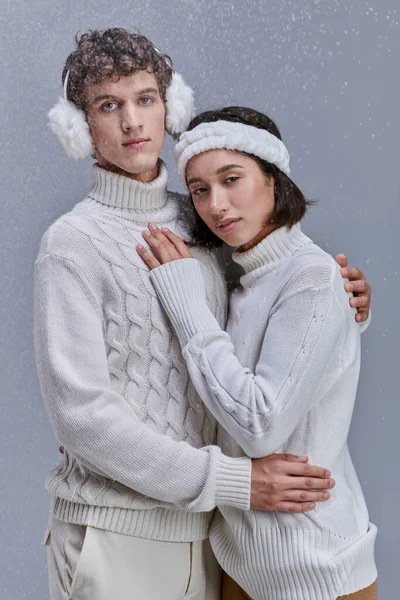 Coppia interrazziale in maglioni bianchi che si abbracciano sotto la neve che cade su sfondo grigio, moda invernale — Foto stock
