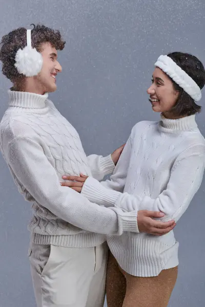 Couple interracial gai en tenue d'hiver confortable se regardant sur fond gris avec de la neige — Photo de stock