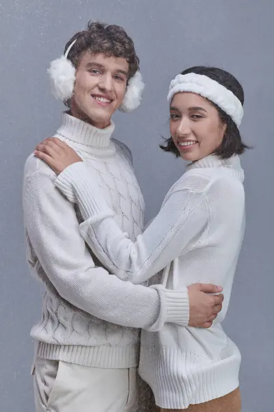 Trendy interracial Paar umarmt und lächelnd Kamera auf grauem Hintergrund mit Schnee, Winter-Stil — Stockfoto