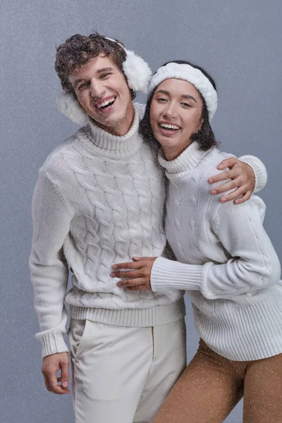 Схвильована міжрасова пара в теплих светрах, що приймають і сміються під падаючим снігом, щаслива зима — стокове фото