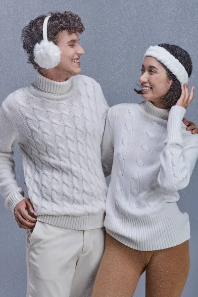 Zufriedenes multiethnisches Paar in weißen Pullovern, die einander auf grauem Hintergrund mit Schnee anschauen — Stockfoto