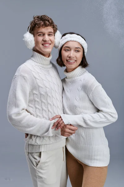 Sonriente pareja interracial en acogedores suéteres de punto posando sobre fondo gris con nieve, invierno de moda - foto de stock