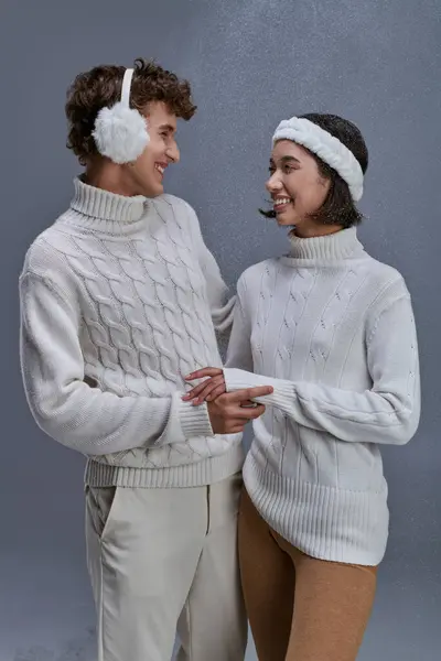 Happy interracial couple in sweaters looking at each other on grey snowy backdrop, romantic winter — Stock Photo