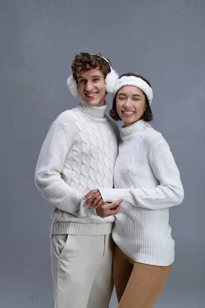 Alegre pareja multiétnica en suéteres sonriendo bajo la nieve cayendo sobre fondo gris, invierno de moda - foto de stock