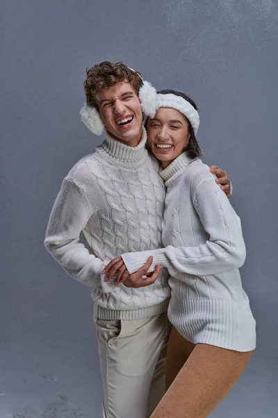 Pareja interracial en suéteres blancos abrazando y riendo sobre fondo gris nevado, moda de invierno - foto de stock