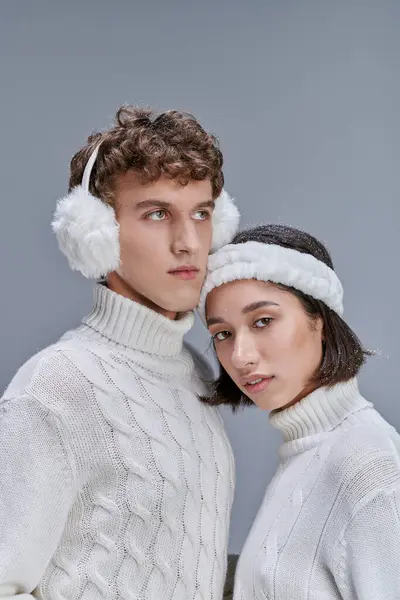 Giovane donna asiatica con i capelli innevati guardando la fotocamera vicino all'uomo in calde cuffie su grigio, stile invernale — Foto stock