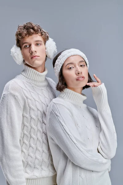 Young and stylish multiethnic couple in white cozy sweaters posing with snowy hair on grey backdrop — Stock Photo