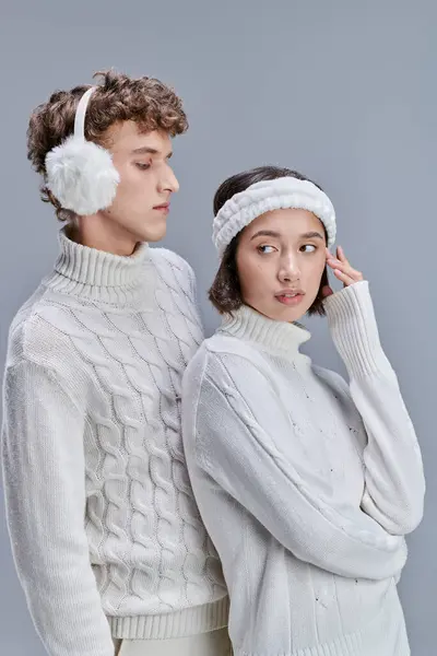 Stylische asiatische Frau, die in der Nähe eines jungen Mannes mit warmen Ohrenschützern auf grauem Hintergrund wegschaut, schneebedecktes Haar — Stockfoto