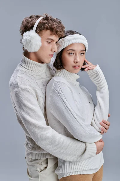 Junger Mann mit Ohrenschützer umarmt charmante asiatische Frau auf grauem Hintergrund, romantisches Paar im Winter — Stockfoto