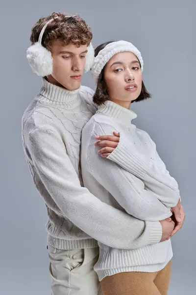 Trendy man in earmuffs hugging young asian woman on grey background, stylish romantic winter — Stock Photo