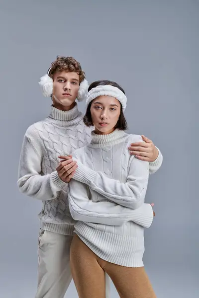 Mann mit Winterohrenschützer umarmt trendige asiatische Frau im Pullover auf grauem, modischem Lookbook-Konzept — Stockfoto