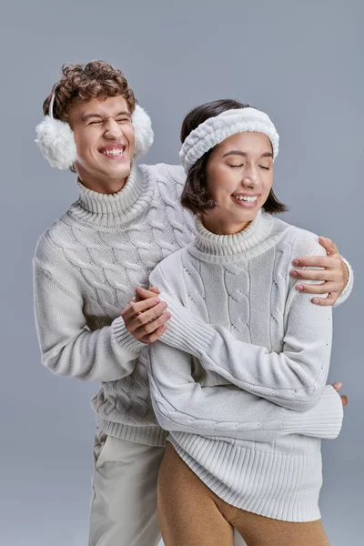 Overjoyed man in cozy earmuffs hugging asian woman in winter sweater on grey, seasonal style — Stock Photo