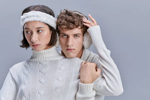 Charming woman touching snowy hair of man in warm earmuffs on grey background, coxy winter style — Stock Photo
