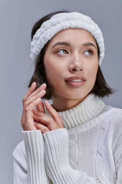 Мечтательная азиатка в трикотажном свитере и мягком повязке, смотрящая в сторону серого, современного зимнего тренда — стоковое фото