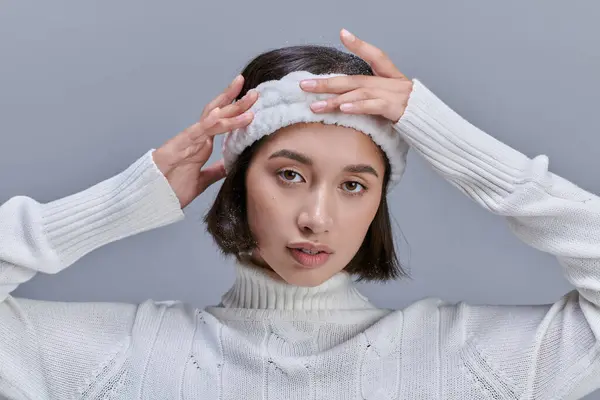 Giovane donna asiatica in maglione bianco regolazione fascia morbida sul grigio, concetto di moda invernale — Foto stock