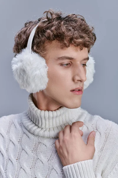 Modischer Mann mit schneebedeckten welligen Haaren posiert in Strickpullover und warmen Ohrenschützern vor grauem Hintergrund — Stockfoto