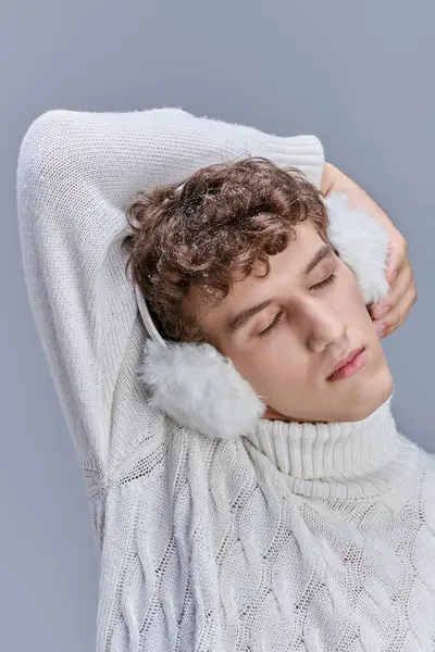 Молодий чоловік в білому в'язаному светрі і вухах позує із закритими очима на сірій, затишній зимовій моді — стокове фото