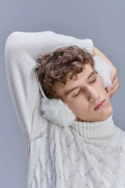 Young man in white knitted sweater and earmuffs posing with closed eyes on grey, cozy winter fashion — Stock Photo