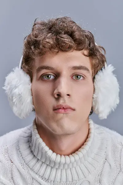 Homme aux cheveux ondulés enneigés et au regard expressif portant des cache-oreilles d'hiver et regardant la caméra sur gris — Photo de stock