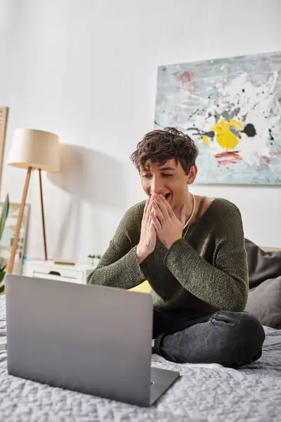 Happy curly transgender blogger using laptop and sitting on bed in modern apartment, social media — Stock Photo