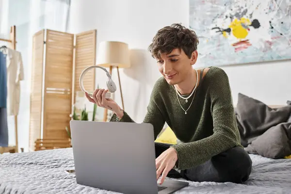 Happy curly transgender blogger using laptop and sitting on bed near smartphone and headphones — Stock Photo