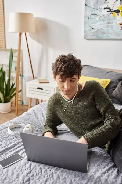 Joven blogger transgénero rizado utilizando el ordenador portátil y sentado en la cama cerca de teléfonos inteligentes y auriculares - foto de stock