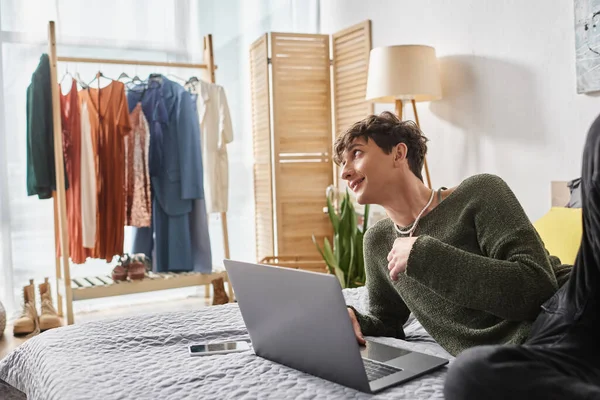 Happy curly transgender blogger using laptop and sitting near smartphone with blank screen on bed — Stock Photo