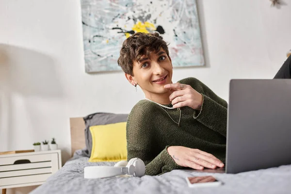 Glücklich lockige Transgender-Bloggerin mit Laptop und auf dem Bett neben Smartphone und Kopfhörer sitzen — Stockfoto