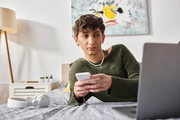 Young curly transgender blogger using smartphone near laptop and sitting on bed near headphones — Stock Photo