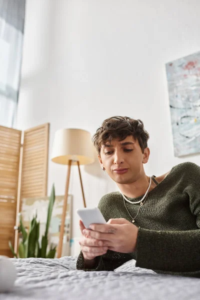 Displeased transgender blogger with curly hair using smartphone in bedroom, social media influencer — Stock Photo