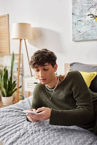 Astonished transgender blogger with curly hair using smartphone in bedroom, social media influencer — Stock Photo