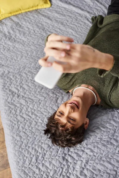 Happy transgender blogger with curly hair using smartphone and lying on bed, social media influencer — Stock Photo