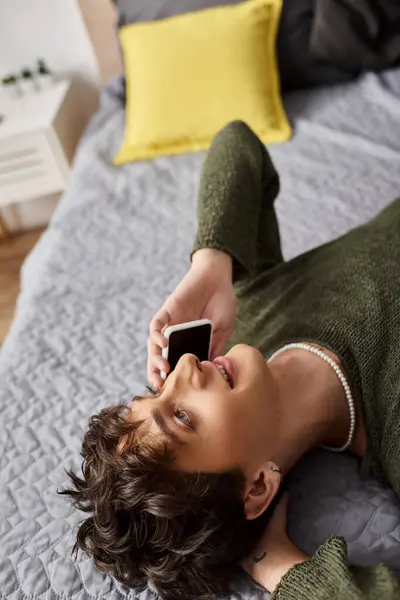 Happy and curly transgender person in casual attire talking on smartphone in bedroom, overhead view — Stock Photo
