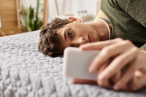 Transgender blogger with curly hair using smartphone and lying on bed, social media influencer — Stock Photo
